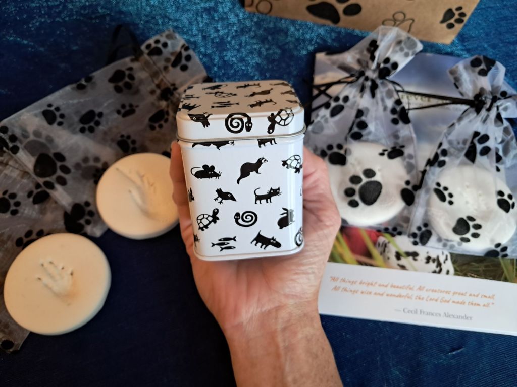A decorative tin with black animal silhouettes held in a hand, surrounded by paw print bags and circular white items in the background.
